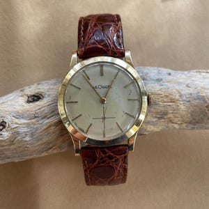 May include: A gold-toned wristwatch with a light-colored face and brown leather band. The watch face has a simple design with thin markers and hands. The band is a rich brown color with a textured pattern. The watch is a classic style.