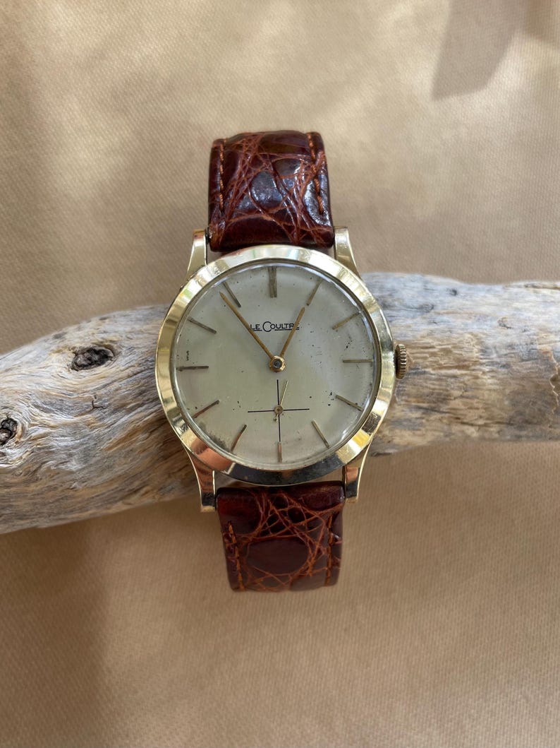 May include: A gold-toned wristwatch with a light-colored face and brown leather band. The watch face has a simple design with thin markers and hands. The band is a rich brown color with a textured pattern. The watch is a classic style.