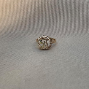 Puede incluir: Un anillo de tono dorado que presenta un marco circular con un borde de estilo cuerda trenzada. En el centro del círculo hay una letra R mayúscula en cursiva. La banda del anillo es delgada y lisa, conectada al elemento decorativo.