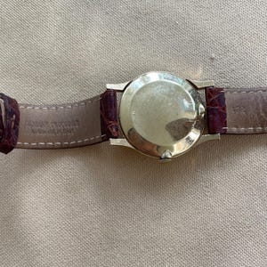 May include: A gold-toned wristwatch with a round face and a brown leather band. The band is made of genuine crocodile leather. The watch is displayed against a neutral-toned background.