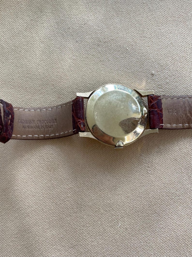 May include: A gold-toned wristwatch with a round face and a brown leather band. The band is made of genuine crocodile leather. The watch is displayed against a neutral-toned background.