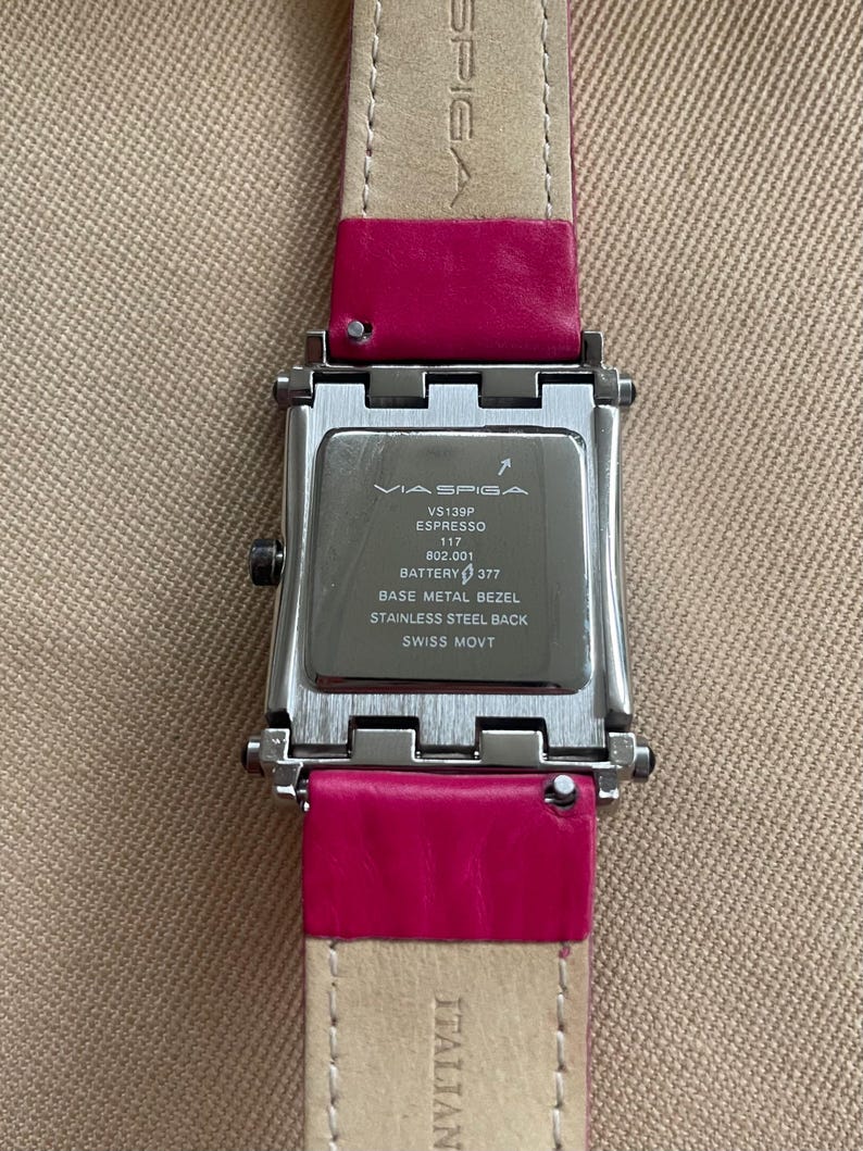 Puede incluir: Un reloj plateado con una correa de cuero rosa. La esfera del reloj es rectangular y tiene un bisel plateado. La esfera del reloj es negra y tiene el texto "VIA SPIGA VS139P ESPRESSO 117 802.001 BATTERY 377 BASE METAL BEZEL STAINLESS STEEL BACK SWISS MOVT ITALIAN" impreso en ella.