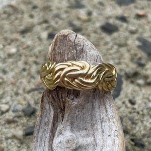 Puede incluir: Un anillo de color dorado con un dise&ntilde;o trenzado. El anillo est&aacute; sentado en un trozo de madera a la deriva.