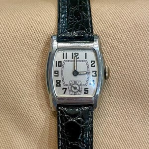 May include: A rectangular silver wristwatch with a white face and black Roman numerals. The watch has a small seconds dial and is attached to a black leather strap.