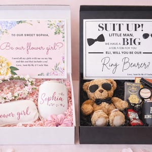 May include: Two gift boxes, one white and one black, with items for a flower girl and ring bearer. The white box includes a floral headband, a personalized tumbler, and a cosmetic bag. The black box contains a teddy bear, sunglasses, and a tumbler.