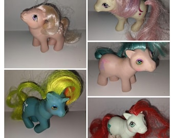 May include: Four vintage My Little Pony toys. The ponies are pink, blue, white, and light blue. They have different colored manes and tails. Each pony has a unique symbol on its flank.