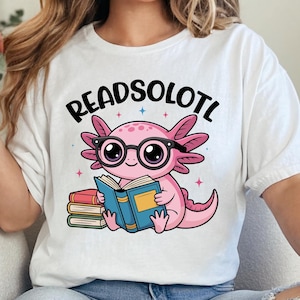 Puede incluir: Camiseta blanca con el texto "READSOLOTL" sobre un ajolote de dibujos animados con gafas y leyendo un libro. El ajolote es rosa, con un libro azul y una pila de libros a la izquierda.