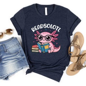 Puede incluir: Camiseta azul marino con un ajolote de dibujos animados con gafas y leyendo un libro. El texto "READSOLOTL" est&aacute; encima del ajolote. Tambi&eacute;n se ven unos pantalones cortos vaqueros y sandalias.