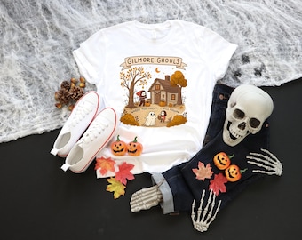 Könnte beinhalten: Weißes T-Shirt mit einem Halloween-Motiv mit der Aufschrift "Gilmore Ghouls, Where You Haunt, I Will Follow". Das Bild zeigt auch weiße Turnschuhe, eine dunkelblaue Jeans, einen Totenkopf sowie dekorative Kürbisse und Blätter.