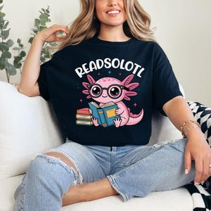 Puede incluir: Camiseta negra con el texto "READSOLOTL" sobre un ajolote de dibujos animados con gafas y leyendo un libro. El ajolote es rosa y el libro es azul. La camiseta la lleva una persona.
