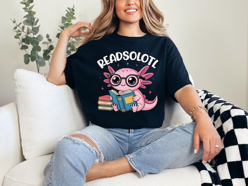 Puede incluir: Camiseta negra con el texto "READSOLOTL" sobre un ajolote de dibujos animados con gafas y leyendo un libro. El ajolote es rosa y el libro es azul. La camiseta la lleva una persona.