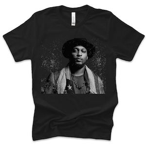 Può includere: T-shirt nero con un ritratto in bianco e nero di una persona con cappello, collana e sciarpa. Il ritratto presenta un motivo a stelle e strisce. La maglietta è realizzata in materiale morbido.