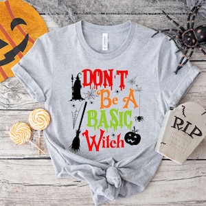 May include: A light gray t-shirt with the text "DON'T Be A BASIC Witch" in red, orange, and green. The design includes a witch silhouette, spiderwebs, a broom, and a jack-o'-lantern. Perfect for Halloween.