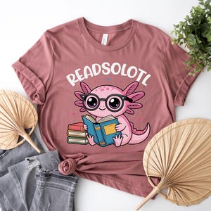 Puede incluir: Camiseta rosa polvoriento con un ajolote de dibujos animados con gafas y leyendo un libro. El texto "READSOLOTL" est&aacute; encima del ajolote. La camiseta se combina con vaqueros grises y un abanico decorativo.