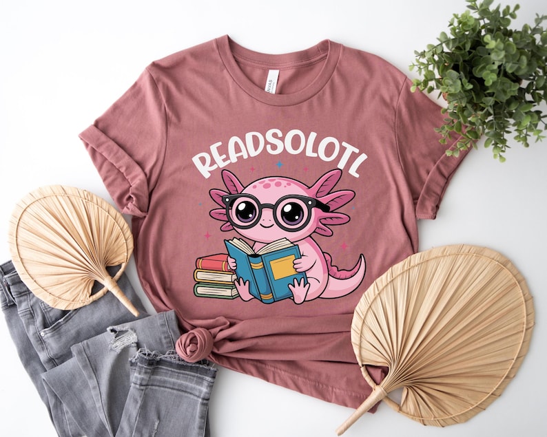Puede incluir: Camiseta rosa polvoriento con un ajolote de dibujos animados con gafas y leyendo un libro. El texto "READSOLOTL" est&aacute; encima del ajolote. La camiseta se combina con vaqueros grises y un abanico decorativo.
