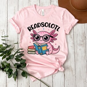 Puede incluir: Camiseta rosa claro con un ajolote de dibujos animados con gafas y leyendo un libro. El texto "READSOLOTL" est&aacute; encima del ajolote. La camiseta tiene las mangas dobladas.