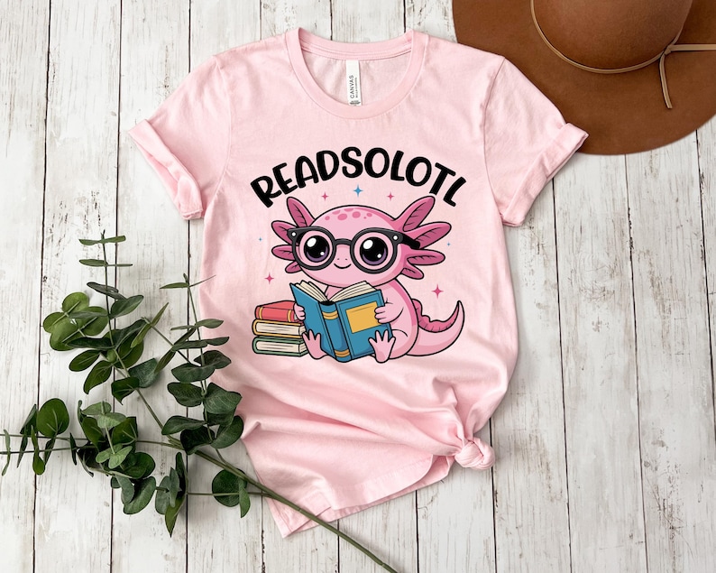 Puede incluir: Camiseta rosa claro con un ajolote de dibujos animados con gafas y leyendo un libro. El texto "READSOLOTL" est&aacute; encima del ajolote. La camiseta tiene las mangas dobladas.