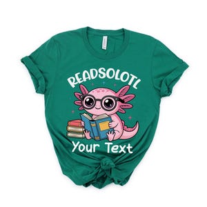 Puede incluir: Una camiseta verde azulado con un ajolote de dibujos animados con gafas y leyendo un libro. El texto "READSOLOTL" y "Your Text" est&aacute;n impresos encima y debajo de la ilustraci&oacute;n del ajolote. El ajolote es rosa y el libro es azul.