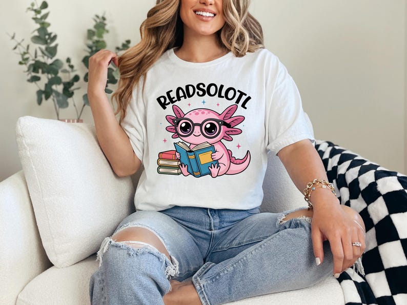 Puede incluir: Camiseta blanca con el texto "READSOLOTL" sobre un ajolote de dibujos animados con gafas y leyendo un libro. El ajolote es rosa, con un libro azul y una pila de libros a la izquierda.
