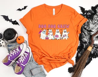 Può includere: T-shirt arancione con la scritta "Boo Boo Crew" e una grafica di fantasmi dei cartoni animati vestiti da infermiere. I fantasmi indossano maschere e trasportano forniture mediche. La maglietta è abbinata a scarpe da ginnastica viola e decorazioni di Halloween.