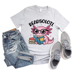 Puede incluir: Camiseta gris claro con el texto "READSOLOTL" sobre un ajolote de dibujos animados con gafas y leyendo un libro. La imagen tambi&eacute;n incluye vaqueros azules y zapatillas grises.