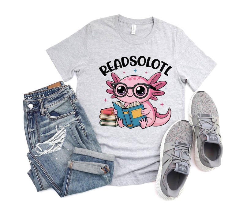 Puede incluir: Camiseta gris claro con el texto "READSOLOTL" sobre un ajolote de dibujos animados con gafas y leyendo un libro. La imagen tambi&eacute;n incluye vaqueros azules y zapatillas grises.