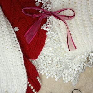 May include: A white knitted scarf with a red knitted scarf tied around it with a pink ribbon. The scarves are decorated with white lace and pearl beads.
