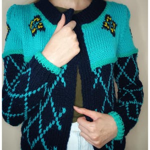 Può includere: Un cardigan di maglia blu e nero con un motivo geometrico. Il cardigan ha una chiusura con bottoni e maniche lunghe.