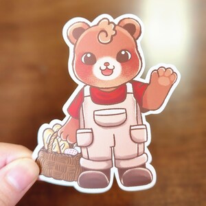 Op de afbeelding: Een sticker met een cartoonbeer die een rood shirt en een beige overall draagt en zwaait. De beer draagt een mand met brood en eieren. De beer heeft een krullend kapsel en een vriendelijke uitdrukking.