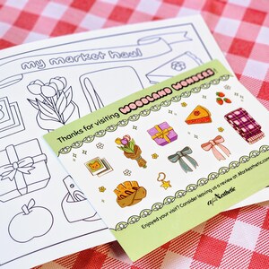 Op de afbeelding: Een groene kaart met de tekst "Thanks for visiting Woodland Wonders" en verschillende illustraties van cadeaus, bloemen en eten. Op de achtergrond een zwart-wit kleurplaat met de tekst "my market haul".