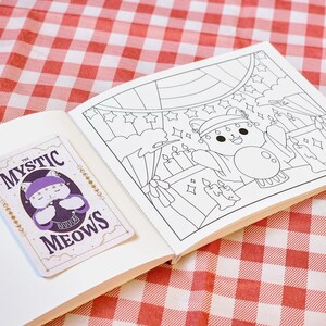 Op de afbeelding: Een open notitieboek met een sticker en een kleurplaat. De sticker toont een paarse kat met de tekst "THE MYSTIC MEOWS". De kleurplaat toont een kattenpersonage in een circusomgeving met kaarsen en sterren. De achtergrond is een rood-wit geblokt patroon.