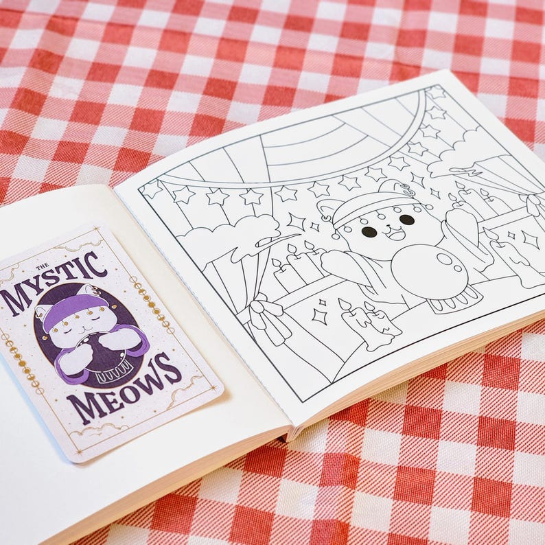 Op de afbeelding: Een open notitieboek met een sticker en een kleurplaat. De sticker toont een paarse kat met de tekst "THE MYSTIC MEOWS". De kleurplaat toont een kattenpersonage in een circusomgeving met kaarsen en sterren. De achtergrond is een rood-wit geblokt patroon.