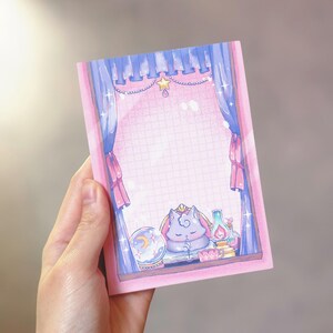 May include: A pink notepad with a whimsical illustration of a sleeping cat in a window. The artwork features blue curtains, a crystal ball, and a lamp. The notepad has a grid pattern and is held by a hand.