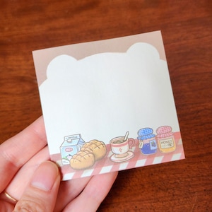 May include: A square sticky note with a bear-shaped top edge. The bottom edge features illustrations of bread, milk, coffee, and jars of jam. The background is white with a red and white checkered pattern.