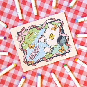 Op de afbeelding: Een vierkante kunstdruk met de woorden "Woodland Wonders" en een cartoonillustratie. De print is omgeven door witte stiften met gekleurde punten, tegen een rood-wit geruite achtergrond.