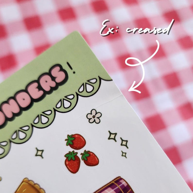 Op de afbeelding: Een vel stickers met een lichtgroene rand en het woord "WONDERS!" in roze en zwart. De stickers bevatten rode aardbeien, een bloem en andere decoratieve elementen. De achtergrond is een rood-wit geblokt patroon.