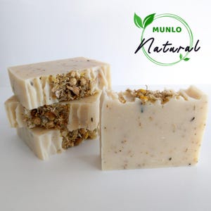 May include: A stack of four white soap bars with dried chamomile flowers embedded in the soap. The soap bars are labelled "MUNLO Natural".