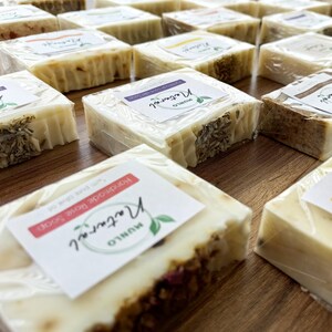 May include: A selection of handmade soap bars, wrapped in plastic. The bars are white and feature various scents and ingredients. Each bar is labelled with the name "Munio" and the scent of the soap. The soaps are displayed on a wooden surface.
