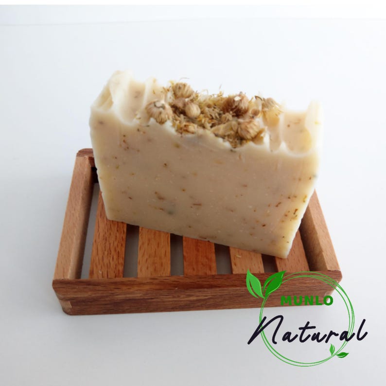 May include: A white bar of soap with dried flowers on top, sitting on a wooden soap dish. The soap dish has the words "Mundo Natural" printed on it.