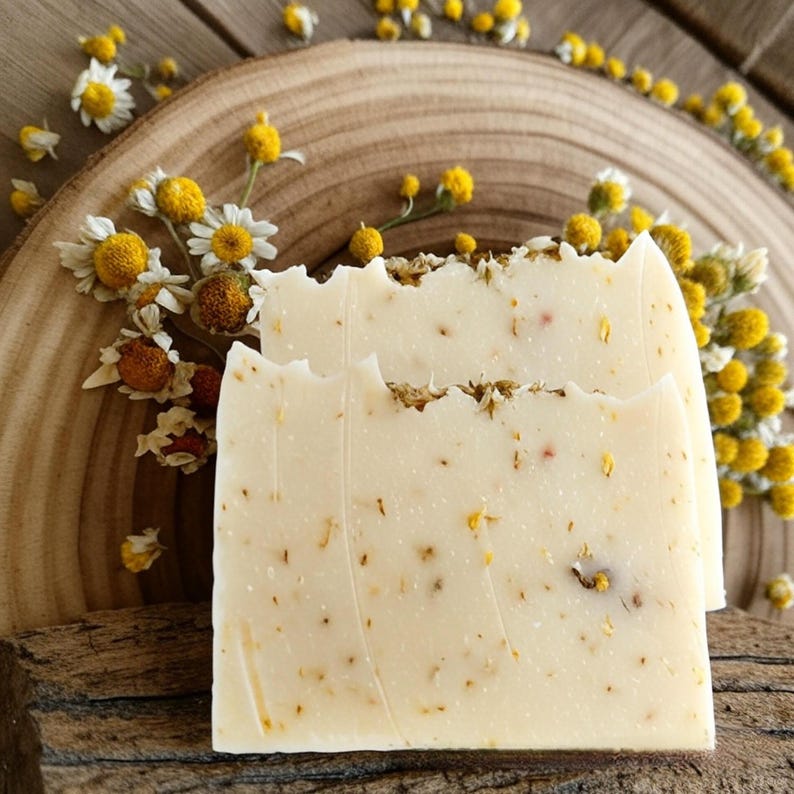 May include: Two bars of white soap with dried yellow flowers embedded in the soap. The soap is sitting on a wooden surface.