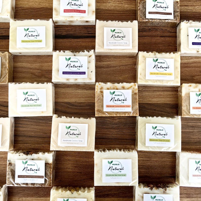 May include: A collection of 16 bars of handmade soap with various scents. The soap bars are arranged in a grid pattern on a wooden surface. Each bar has a label with the words "MUNLO Natural" and the scent of the soap.