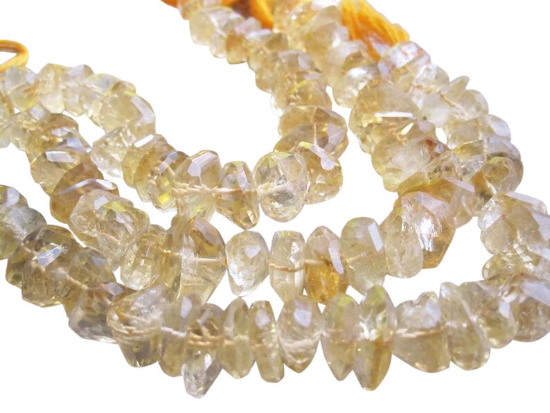 May include: Strands of faceted, light yellow citrine gemstone beads. The beads are irregular in shape and strung on a yellow cord. The gemstones have a translucent quality, reflecting light. The background is white.