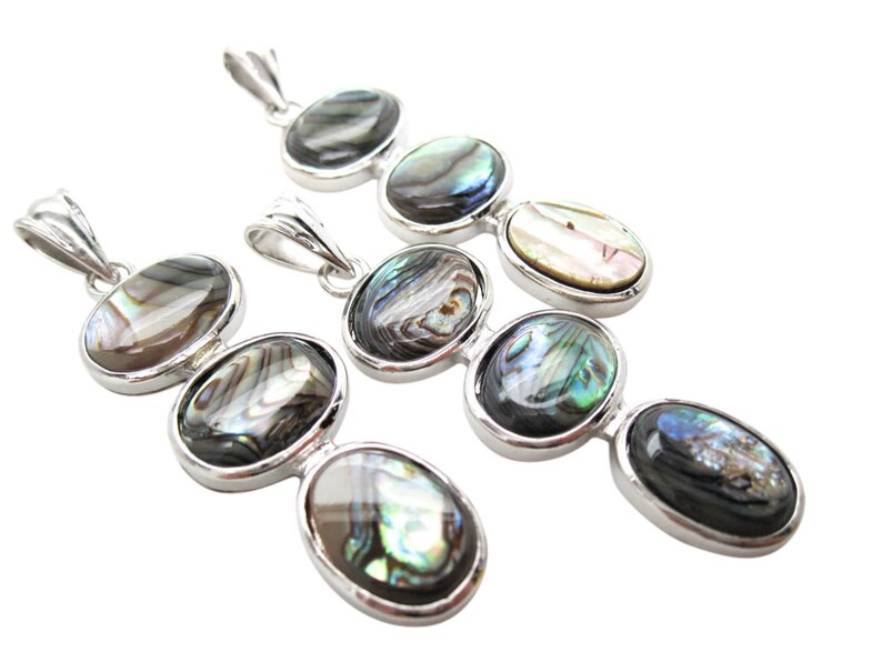 Pode incluir: V&aacute;rios pingentes de prata com incrusta&ccedil;&otilde;es ovais de concha de abalone. Cada pingente apresenta tr&ecirc;s pe&ccedil;as de concha em sequ&ecirc;ncia, com cores iridescentes de verde, azul e branco. Os pingentes s&atilde;o exibidos em fundo branco.