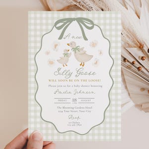 May include: A baby shower invitation with a whimsical design. The invitation features a light green gingham background and a scalloped border. The text reads "Silly Goose" with illustrations of geese and flowers. The invitation is held by a hand.