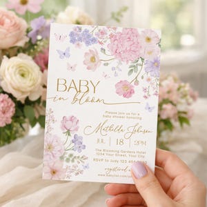 May include: Baby shower invitation featuring floral and butterfly designs in pink, purple, and gold. The card says "Baby in Bloom" and includes event details. The invitation is held in front of a floral arrangement.