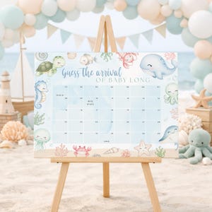 K&ouml;nnte beinhalten: Ein Babyparty-Kalender auf einer h&ouml;lzernen Staffelei mit einem Meeresleben-Thema. Der Kalender ist hellblau mit Illustrationen von Meereskreaturen und dem Text "Guess the arrival of Baby Long". Die Staffelei steht auf sandigem Untergrund.