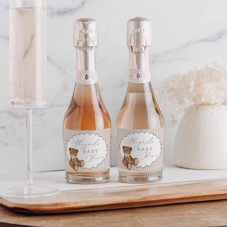 Op de afbeelding: Twee mini-champagneflessen met roze folie doppen en labels met de tekst "Abigail's Baby Shower" met een teddybeer afbeelding. Een champagneglas met roze vloeistof staat links. Een witte vaas met bloemen staat rechts.