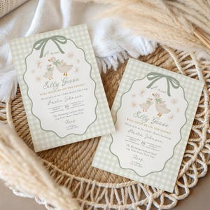 May include: Two baby shower invitations with a light green gingham pattern border. The invitations feature a whimsical illustration of two geese and the text "Silly Goose" and event details. The invitations are placed in a woven basket.