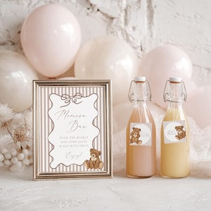 Op de afbeelding: Een ingelijst bord met de tekst "Momosa Bar" en twee glazen flessen met sap, elk met een teddybeer-sticker. Het bord heeft bruine en witte strepen, met een gouden lijst. Roze en witte ballonnen zijn op de achtergrond.
