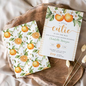 Peut inclure: Deux faire-part de baby shower avec des motifs d'oranges et de fleurs. L'un des faire-part porte le texte "A Little Cutie Is On The Way" et les d&eacute;tails de l'&eacute;v&eacute;nement. L'autre pr&eacute;sente un motif r&eacute;p&eacute;t&eacute; d'oranges, de feuilles et de fleurs.